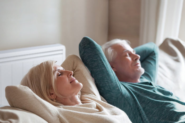 Couple senior dans leur canapé avec un radiateur basse consommation Aterno derrière eux. Le confort thermique est optimal.